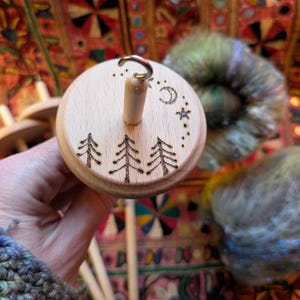 Puede incluir: Un huso de madera con un dise&ntilde;o de bosque y celestial. La parte superior del huso presenta tres &aacute;rboles grabados, una luna creciente y estrellas. El huso se sostiene en una mano, con hilo colorido en el fondo.