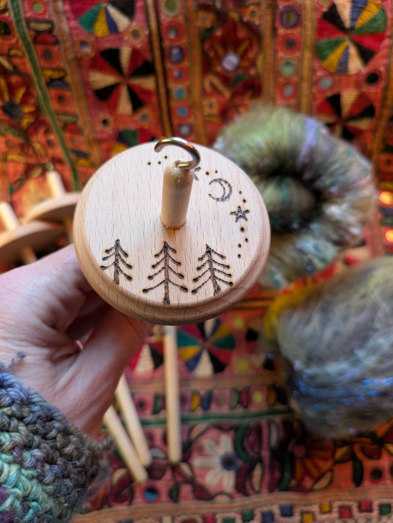 Puede incluir: Un huso de madera con un dise&ntilde;o de bosque y celestial. La parte superior del huso presenta tres &aacute;rboles grabados, una luna creciente y estrellas. El huso se sostiene en una mano, con hilo colorido en el fondo.