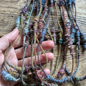 May include: A collection of colorful, handmade fiber art strands in shades of blue, purple, gold, and green. The strands are held in a hand, showcasing the intricate textures and details of the yarn. The art appears to be made of wrapped and knotted threads.