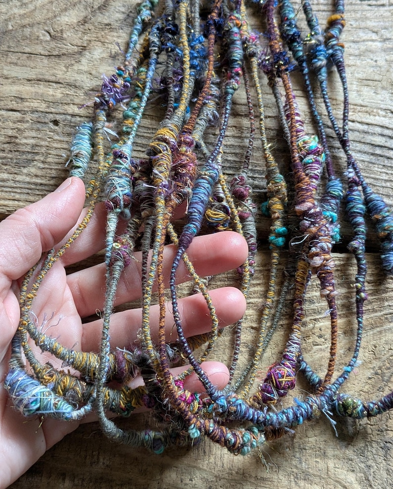 May include: A collection of colorful, handmade fiber art strands in shades of blue, purple, gold, and green. The strands are held in a hand, showcasing the intricate textures and details of the yarn. The art appears to be made of wrapped and knotted threads.