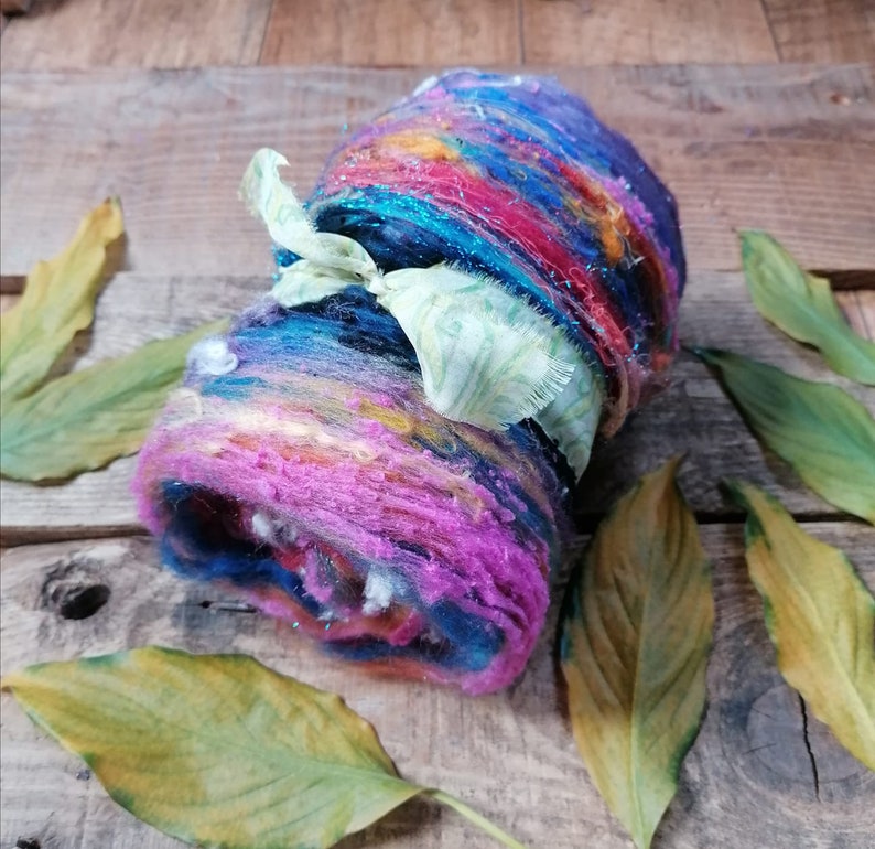May include: A colorful roll of yarn with a green ribbon tied around it. The yarn is a mix of pink, blue, yellow, and orange. The yarn is on a wooden surface with leaves.