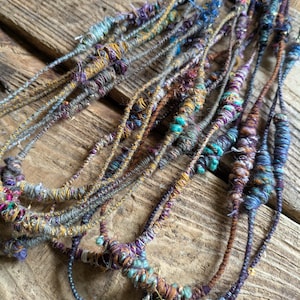 May include: Close-up of several strands of handmade yarn, featuring a variety of colors including teal, purple, gold, and brown. The yarn appears to be textured and knotted, with a rustic, bohemian aesthetic. The strands are arranged on a weathered wooden surface.