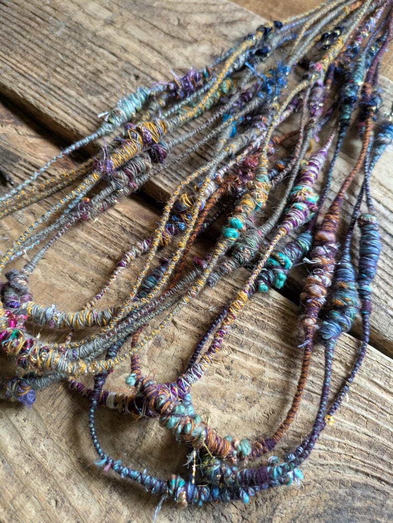 May include: Close-up of several strands of handmade yarn, featuring a variety of colors including teal, purple, gold, and brown. The yarn appears to be textured and knotted, with a rustic, bohemian aesthetic. The strands are arranged on a weathered wooden surface.
