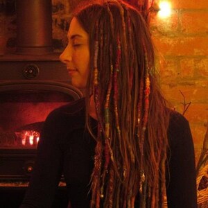 May include: A woman with long, brown dreadlocks with colorful yarn woven in. She is wearing a dark, long-sleeved shirt and is standing in front of a fireplace.