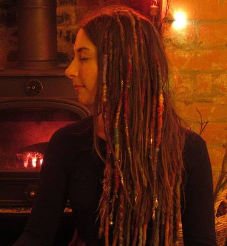 May include: A woman with long, brown dreadlocks with colorful yarn woven in. She is wearing a dark, long-sleeved shirt and is standing in front of a fireplace.