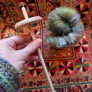Puede incluir: Un huso de madera con una roldana redonda y un gancho en la parte superior, sostenido en una mano. Al lado hay una madeja de hilo verde, azul y morado. El fondo es un textil colorido con patrones bordados.