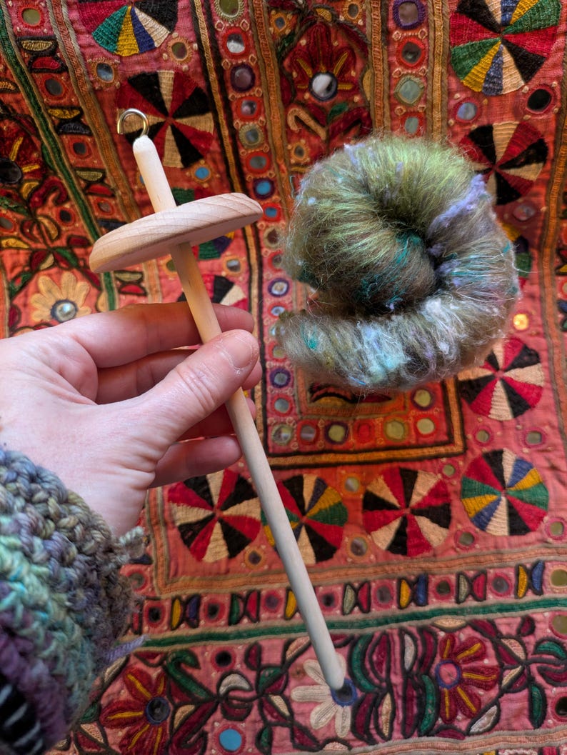 Puede incluir: Un huso de madera con una roldana redonda y un gancho en la parte superior, sostenido en una mano. Al lado hay una madeja de hilo verde, azul y morado. El fondo es un textil colorido con patrones bordados.