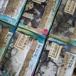 May include: A collection of handmade paper art pieces. Each piece features a unique blend of textures and colors, with visible ink patterns and Japanese text. The pieces are arranged on a wooden surface.