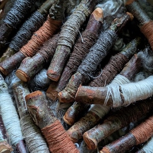 Pode incluir: Uma coleção de paus de madeira envoltos em fios de várias cores, incluindo laranja, cinza e marrom. Os paus têm uma aparência natural e rústica, com casca e nós visíveis. Os fios são enrolados firmemente em volta dos paus, criando uma superfície texturizada. Os paus estão dispostos numa cesta.