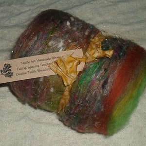 May include: A colorful ball of wool yarn with a brown, green, red, and white color scheme. The yarn is tied with a gold ribbon and a tag that reads "Textile Art, Handmade Goods, Felting, Spinning Supplies, Creative Textile Workshop".