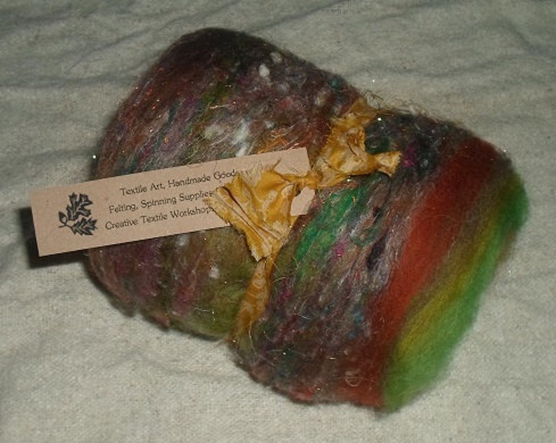 May include: A colorful ball of wool yarn with a brown, green, red, and white color scheme. The yarn is tied with a gold ribbon and a tag that reads "Textile Art, Handmade Goods, Felting, Spinning Supplies, Creative Textile Workshop".