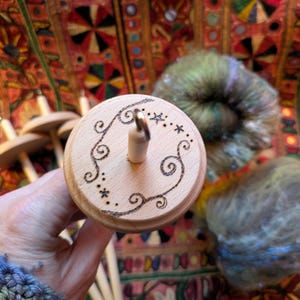 Puede incluir: Un huso de madera con un dise&ntilde;o decorativo de espirales y estrellas grabadas en la parte superior. El huso se sostiene en una mano, con lana colorida en el fondo. Se utiliza para hilar lana a mano.