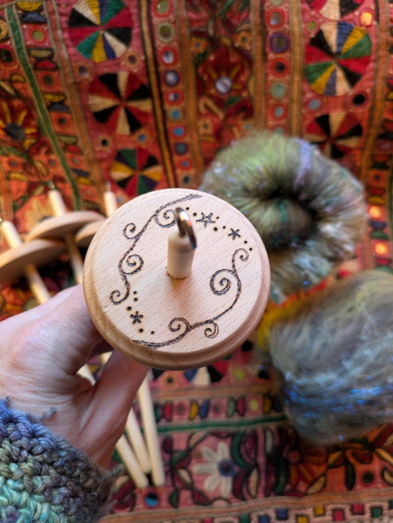Puede incluir: Un huso de madera con un dise&ntilde;o decorativo de espirales y estrellas grabadas en la parte superior. El huso se sostiene en una mano, con lana colorida en el fondo. Se utiliza para hilar lana a mano.