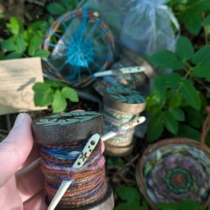 May include: A dark wooden spool wrapped in multicoloured yarn, featuring a white bone needle with a star engraving. The spool top has a green leaf pattern. Additional spools, a small woven basket, and a dreamcatcher are visible in the background.