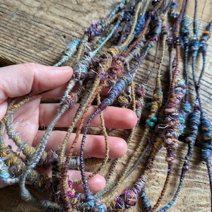 May include: Close-up of several strands of handmade fiber art. The strands are a mix of colors including blue, purple, gold, and brown. The strands are wrapped with various textures and embellishments, creating a unique and artistic look.