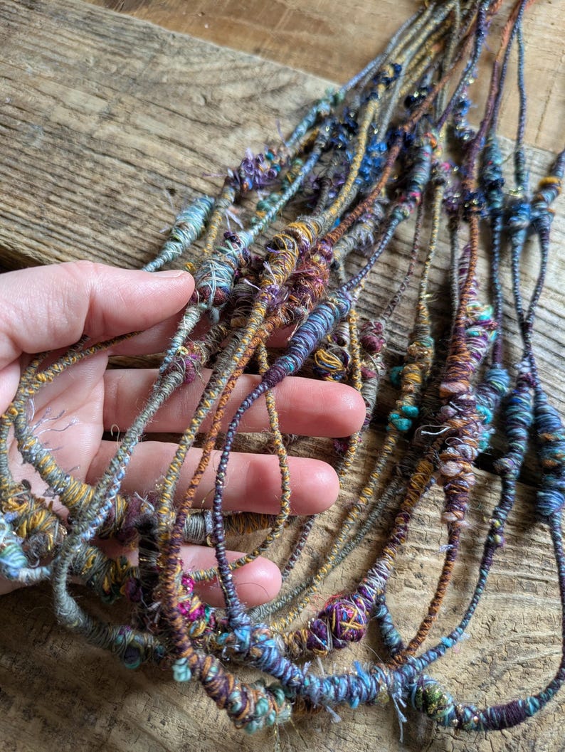 May include: Close-up of several strands of handmade fiber art. The strands are a mix of colors including blue, purple, gold, and brown. The strands are wrapped with various textures and embellishments, creating a unique and artistic look.