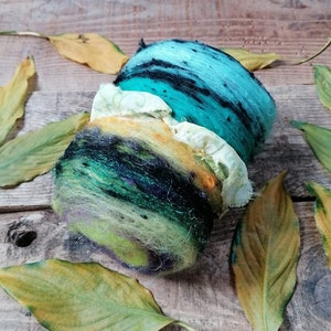 May include: A hand-felted wool ball with a layered design in shades of green, blue, yellow, and black. The ball is surrounded by dried leaves.