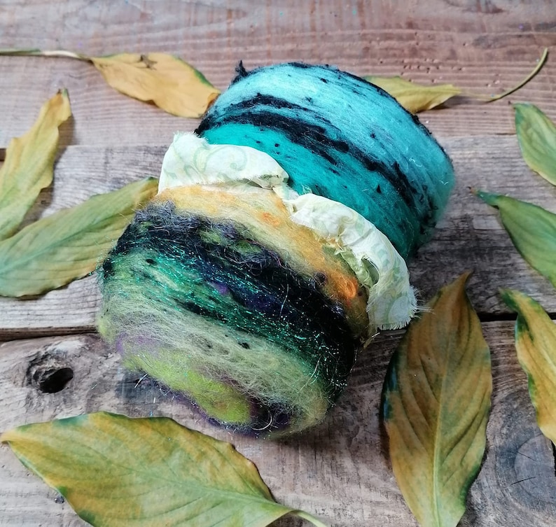 May include: A hand-felted wool ball with a layered design in shades of green, blue, yellow, and black. The ball is surrounded by dried leaves.