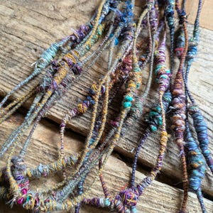 May include: Close-up of several strands of handmade yarn, each intricately wrapped with colorful threads. The yarn features a mix of blues, purples, yellows, and greens, creating a textured and vibrant appearance. The strands are arranged on a rustic wooden surface.