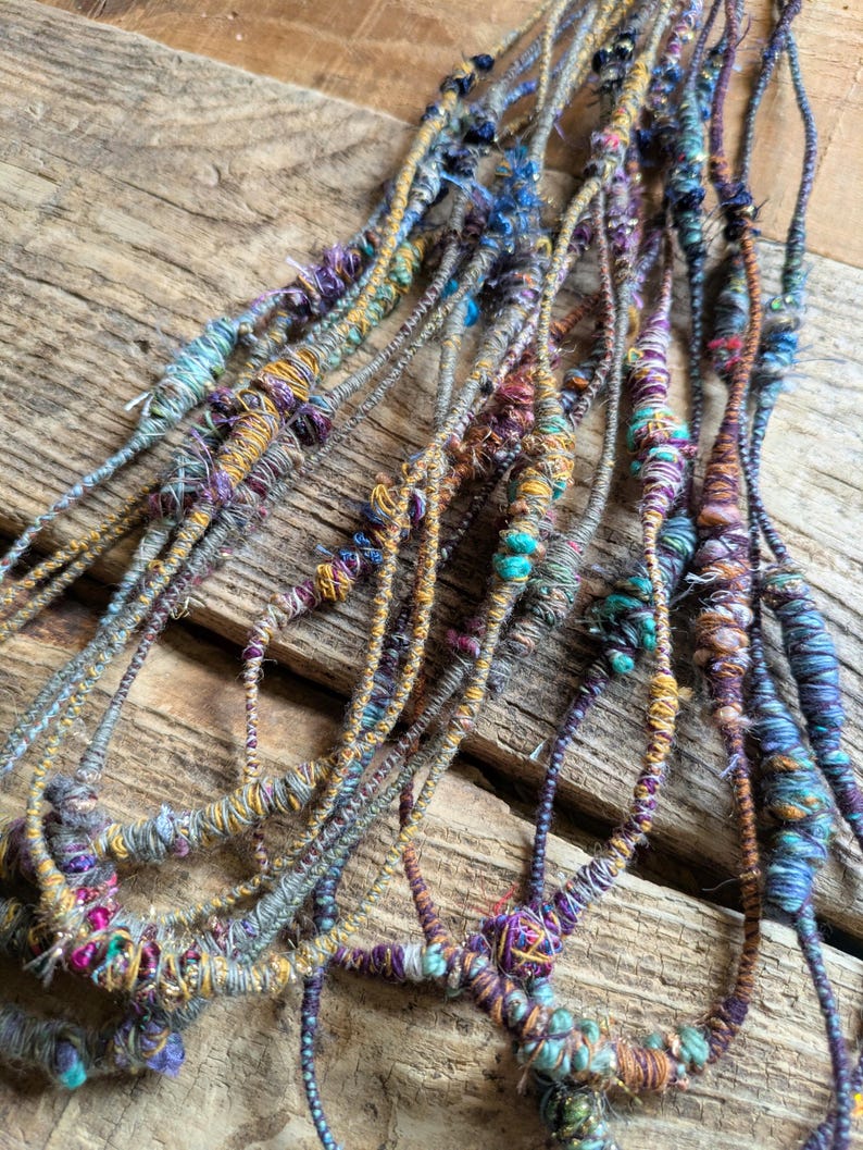 May include: Close-up of several strands of handmade yarn, each intricately wrapped with colorful threads. The yarn features a mix of blues, purples, yellows, and greens, creating a textured and vibrant appearance. The strands are arranged on a rustic wooden surface.