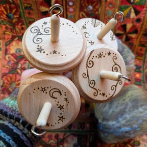 Puede incluir: Cuatro husos de madera con dise&ntilde;os decorativos. Cada huso tiene una base circular de madera con una clavija central de madera y un gancho de metal. Las bases est&aacute;n decoradas con patrones celestiales y arremolinados grabados. Se ve un ovillo de hilo colorido.