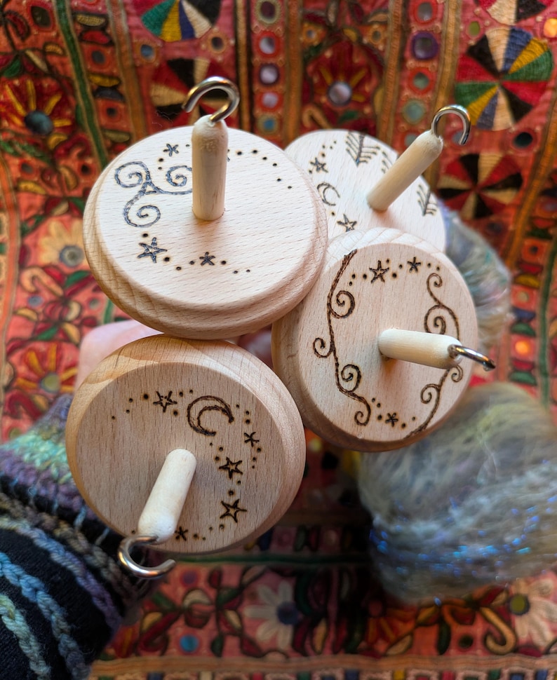 Puede incluir: Cuatro husos de madera con dise&ntilde;os decorativos. Cada huso tiene una base circular de madera con una clavija central de madera y un gancho de metal. Las bases est&aacute;n decoradas con patrones celestiales y arremolinados grabados. Se ve un ovillo de hilo colorido.