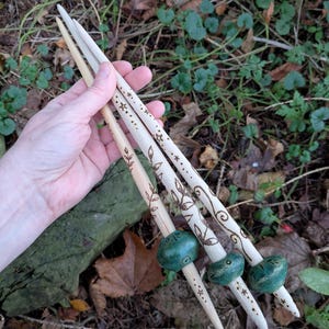 May include: Three wooden hair sticks with decorative designs and green beads. The sticks feature etched patterns of stars, leaves, and swirls. The background is a natural setting with green plants and brown leaves.