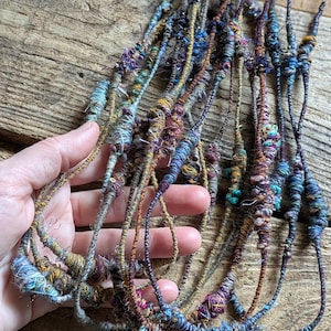 May include: A collection of handmade necklaces with a bohemian aesthetic. The necklaces feature intricate designs with various colors, including blue, purple, and gold. The textured, woven appearance suggests a focus on natural materials and artisanal craftsmanship.