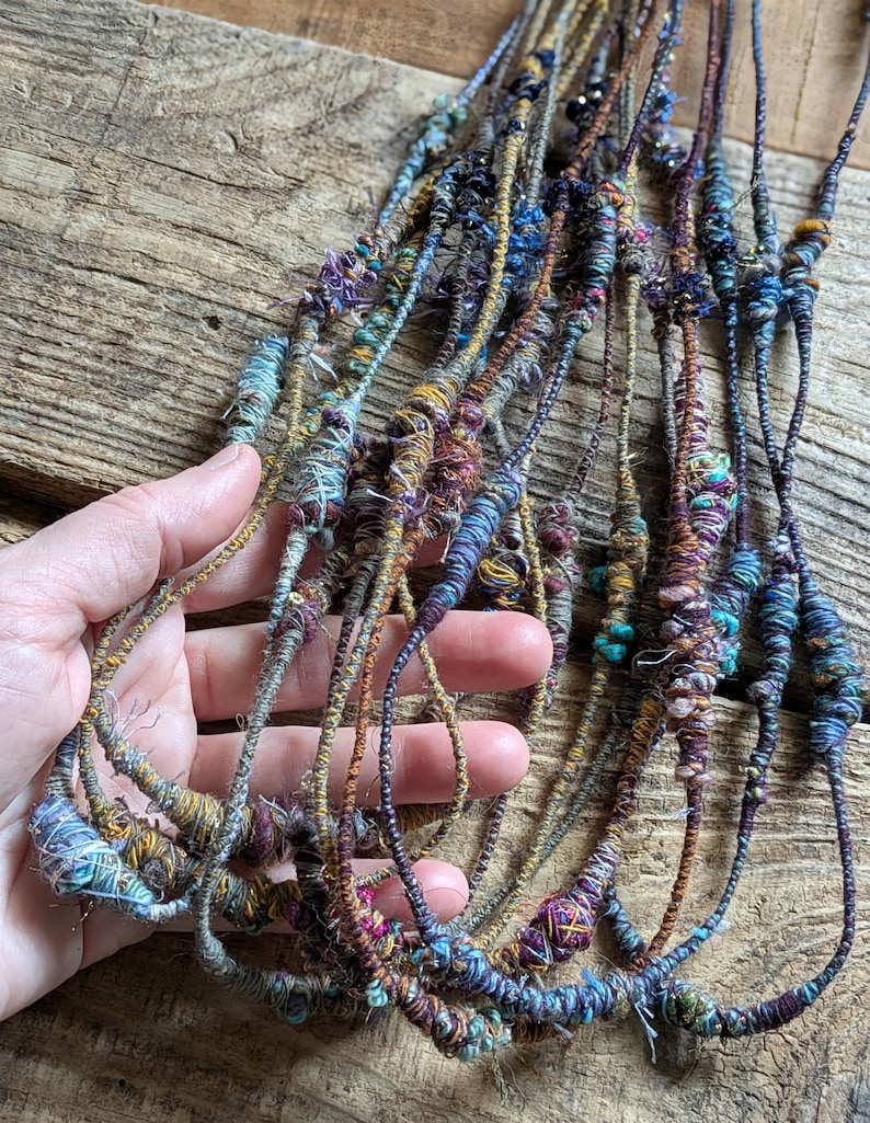 May include: A collection of handmade necklaces with a bohemian aesthetic. The necklaces feature intricate designs with various colors, including blue, purple, and gold. The textured, woven appearance suggests a focus on natural materials and artisanal craftsmanship.