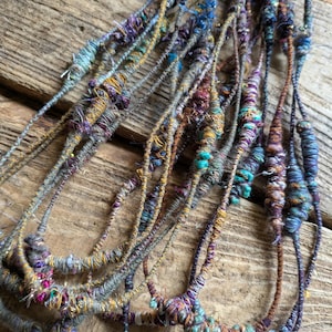 May include: Close-up of several strands of handmade fiber art. The strands are a mix of colors, including teal, purple, gold, and gray. The fiber art appears to be made of yarn and other materials, with a textured and organic look.