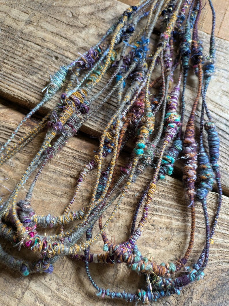 May include: Close-up of several strands of handmade fiber art. The strands are a mix of colors, including teal, purple, gold, and gray. The fiber art appears to be made of yarn and other materials, with a textured and organic look.