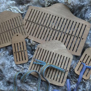 Pode incluir: Um conjunto de ferramentas de tecelagem de madeira, incluindo pentes de tecelagem de vários tamanhos e uma pequena lançadeira, sobre lã cinzenta. As ferramentas são feitas de madeira clara e têm ranhuras verticais.