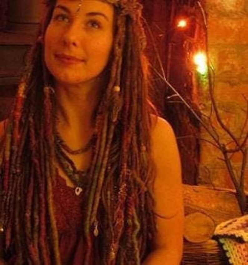 May include: A woman with long, brown dreadlocks decorated with beads and charms. She is wearing a red top and a beaded necklace. The background is a warm, dimly lit room with a brick wall and string lights.
