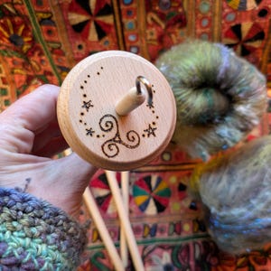 Puede incluir: Un huso de madera con un dise&ntilde;o decorativo de estrellas y remolinos. El huso se sostiene en una mano, con lana colorida en el fondo. El huso es una herramienta para hilar lana a mano.