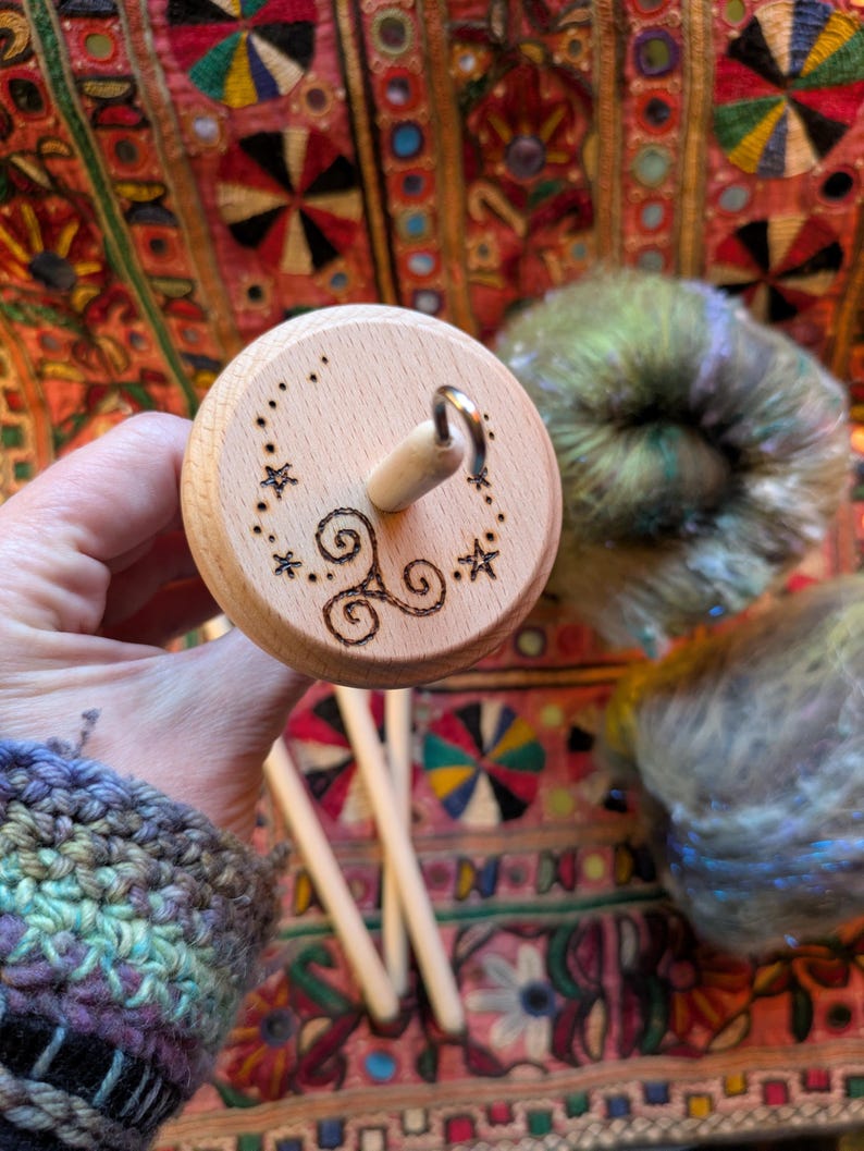 Puede incluir: Un huso de madera con un dise&ntilde;o decorativo de estrellas y remolinos. El huso se sostiene en una mano, con lana colorida en el fondo. El huso es una herramienta para hilar lana a mano.