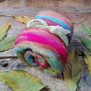 May include: A colorful roll of wool fiber with shades of pink, orange, yellow, blue, and green. The wool is tied with a white ribbon and is sitting on a wooden surface with dried leaves.
