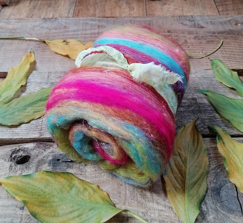 May include: A colorful roll of wool fiber with shades of pink, orange, yellow, blue, and green. The wool is tied with a white ribbon and is sitting on a wooden surface with dried leaves.