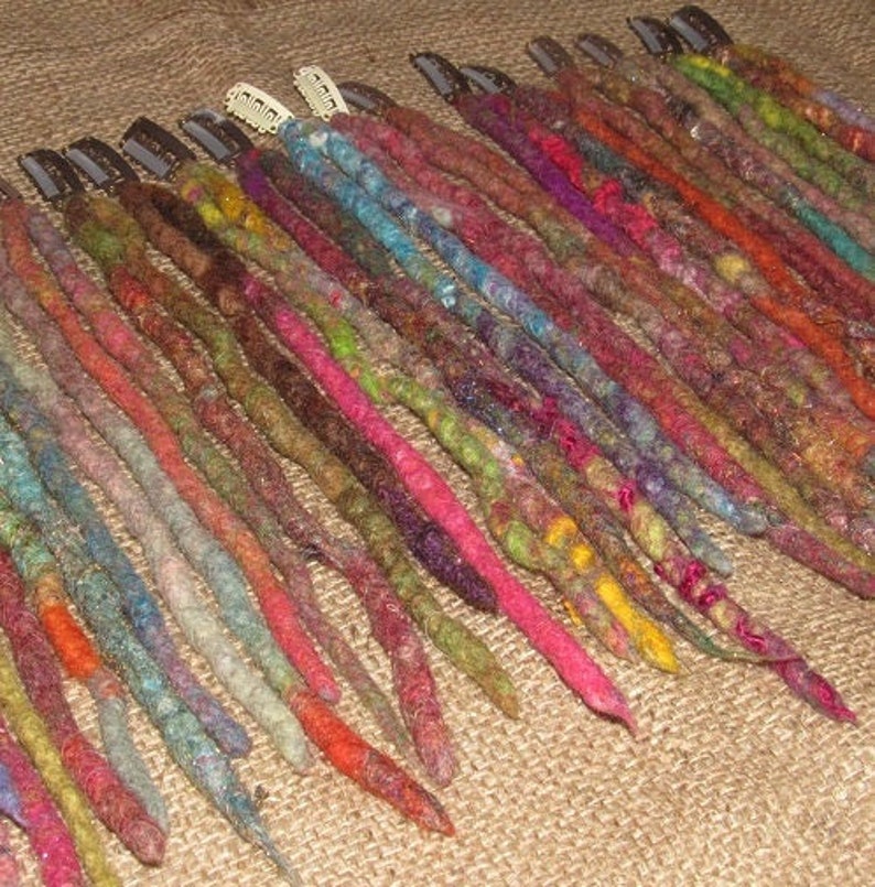 May include: A collection of colorful, multi-colored, and textured dreadlock extensions. The extensions are arranged in a row and are secured with small clips.