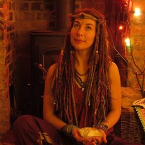 May include: A woman with long, colorful dreadlocks wears a beaded headband and a layered necklace. She is sitting in front of a brick fireplace with a wood-burning stove. The woman is holding a white stone in her hands.