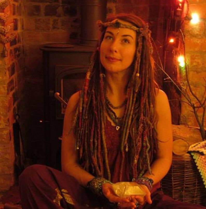 May include: A woman with long, colorful dreadlocks wears a beaded headband and a layered necklace. She is sitting in front of a brick fireplace with a wood-burning stove. The woman is holding a white stone in her hands.