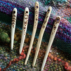 May include: Five wooden shawl pins with decorative burnt designs. Each pin features a unique pattern, including leaves, a crescent moon, stars, and a sun. The pins are light-coloured and rest on a colourful, textured textile.