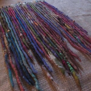 May include: A collection of colorful wool dreadlock extensions. The dreadlocks are arranged in rows and are various shades of blue, green, purple, red, and brown. Some of the dreadlocks have beads or other embellishments.
