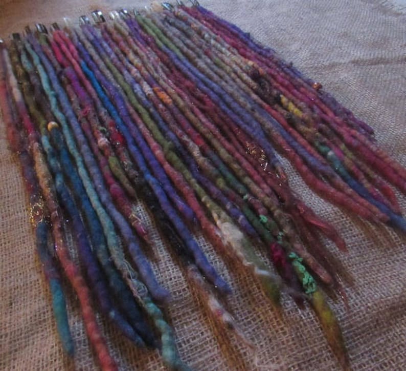 May include: A collection of colorful wool dreadlock extensions. The dreadlocks are arranged in rows and are various shades of blue, green, purple, red, and brown. Some of the dreadlocks have beads or other embellishments.