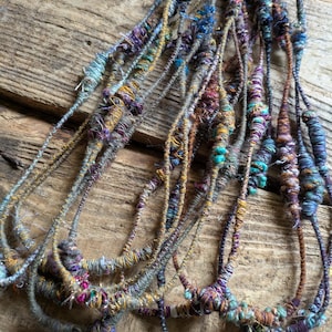May include: A collection of handmade fiber art necklaces. The necklaces feature a variety of colors, including teal, purple, gold, and gray. The textures are varied, with some sections appearing thicker and more textured than others. The necklaces are displayed on a wooden surface.
