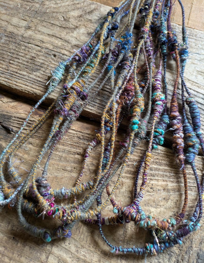 May include: A collection of handmade fiber art necklaces. The necklaces feature a variety of colors, including teal, purple, gold, and gray. The textures are varied, with some sections appearing thicker and more textured than others. The necklaces are displayed on a wooden surface.