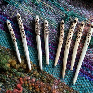 Könnte beinhalten: Eine Sammlung handgefertigter Haarstäbe aus Holz. Die Stäbe sind hellfarbig mit dekorativen, dunkelbraunen geätzten Designs, darunter Wirbel, Blätter und Sterne. Die Haaraccessoires werden auf einem farbenfrohen, strukturierten Textilhintergrund präsentiert.