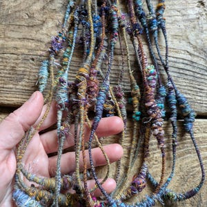May include: A collection of colorful, handmade fiber art strands. The strands are a mix of blues, purples, greens, and golds, with a textured, woven appearance. The strands are held in a hand, showcasing the intricate details and varied textures.