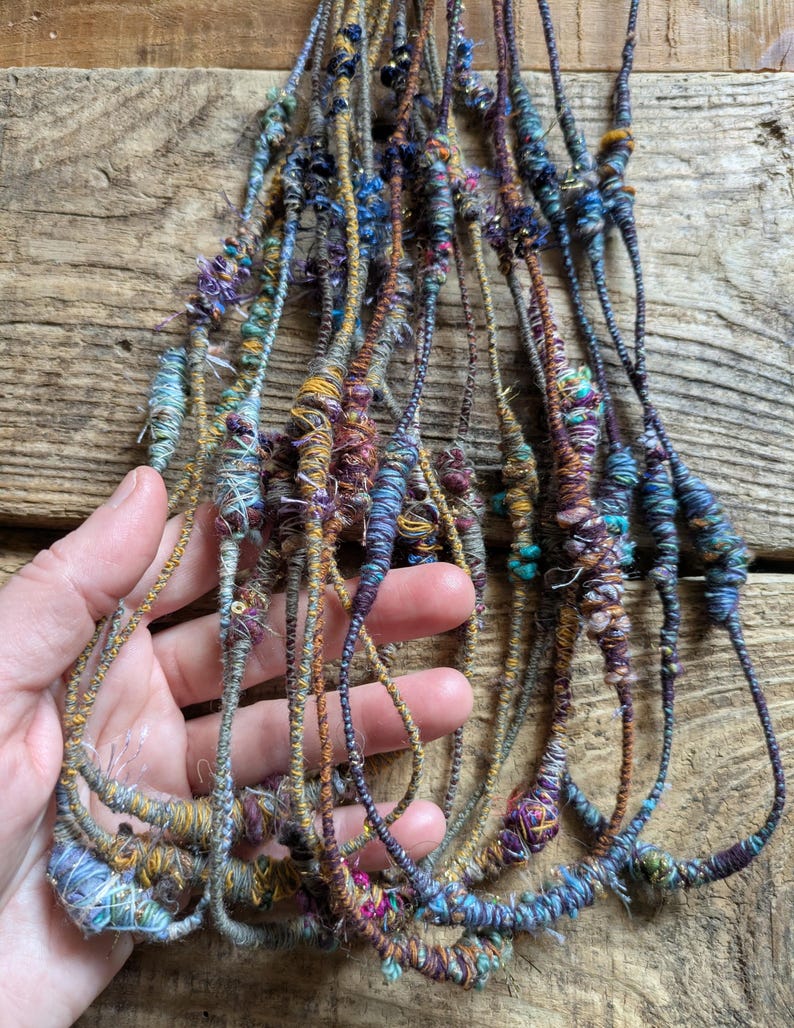 May include: A collection of colorful, handmade fiber art strands. The strands are a mix of blues, purples, greens, and golds, with a textured, woven appearance. The strands are held in a hand, showcasing the intricate details and varied textures.