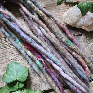 May include: A set of colourful wool dreadlocks in shades of purple, blue, brown, and pink. The dreadlocks are arranged on a wooden surface with green leaves and a crystal.