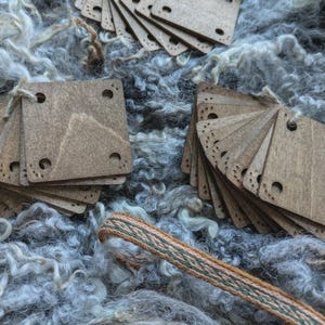May include: Multiple stacks of square wooden tags with rounded corners and small holes are arranged on grey wool. Each tag has a series of small, decorative holes along one edge. A woven cord with green and brown stripes is also present.